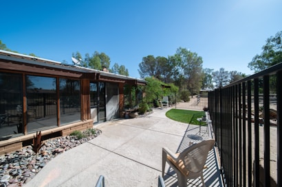 An outdoor patio area adjacent to a house with large windows. The patio is paved with concrete and has a section with decorative stones and plants. There is a metal fence running along the right side, and a plastic chair is placed near the fence. Trees are visible in the background against a clear blue sky.