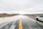 A flooded road stretches ahead under a cloudy sky. Water covers parts of the asphalt, creating a reflective surface. A white, scratched car is parked at the side of the road. The surrounding landscape is barren with dry vegetation and distant hills.