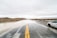 A flooded road stretches ahead under a cloudy sky. Water covers parts of the asphalt, creating a reflective surface. A white, scratched car is parked at the side of the road. The surrounding landscape is barren with dry vegetation and distant hills.