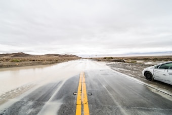 A flooded road stretches ahead under a cloudy sky. Water covers parts of the asphalt, creating a reflective surface. A white, scratched car is parked at the side of the road. The surrounding landscape is barren with dry vegetation and distant hills.