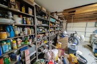A cluttered storage area with shelves filled with various items such as cleaning products, cans, and boxes. On the floor are stacked cardboard boxes, plastic bins, and a bicycle. The ceiling is partially open, showing wooden beams. A fan is also visible among the items.