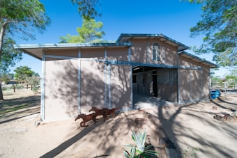 A tan-colored stable with a high roof set amidst a rural area. Shadows of trees are cast on the building's walls, and there are plants and shadows on the sandy ground. Metal railings and a small window are features of the stable. A piece of outdoor decor shaped like horses is visible in front of the structure.