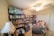 A well-stocked storage room featuring numerous shelves filled with canned goods, household supplies, and essential items. There are several packs of paper towels stacked on the top shelf, and various food products, including canned vegetables and sauces, occupying most of the other shelves. A table lamp is positioned near a chair in the foreground, and a ceiling fan is visible on the ceiling.