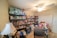 A well-stocked storage room featuring numerous shelves filled with canned goods, household supplies, and essential items. There are several packs of paper towels stacked on the top shelf, and various food products, including canned vegetables and sauces, occupying most of the other shelves. A table lamp is positioned near a chair in the foreground, and a ceiling fan is visible on the ceiling.