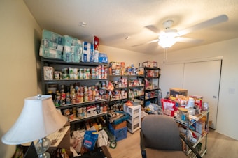 A well-stocked storage room featuring numerous shelves filled with canned goods, household supplies, and essential items. There are several packs of paper towels stacked on the top shelf, and various food products, including canned vegetables and sauces, occupying most of the other shelves. A table lamp is positioned near a chair in the foreground, and a ceiling fan is visible on the ceiling.