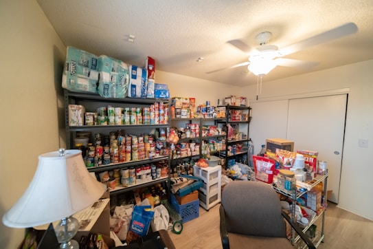 A well-stocked storage room featuring numerous shelves filled with canned goods, household supplies, and essential items. There are several packs of paper towels stacked on the top shelf, and various food products, including canned vegetables and sauces, occupying most of the other shelves. A table lamp is positioned near a chair in the foreground, and a ceiling fan is visible on the ceiling.