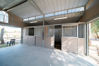 This image depicts the interior of a clean, well-maintained building with multiple subdivided compartments, suggestive of a stable or animal housing area. The compartments have metal bars on the upper sections and solid lower sections, with an open space leading outside visible through the structures. Natural light is coming in through large windows, and a black bench is positioned near the entrance.