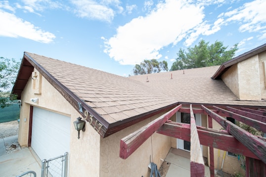 A residential house with a beige exterior and a brown shingle roof. The roof features several chimneys, and there's a white garage door on the side. A decorative lamp is mounted near the garage. Red wooden beams are visible near the entrance, possibly part of a pergola structure. The house number '9347' is displayed prominently. Bushes and trees are seen in the background under a partly cloudy sky.