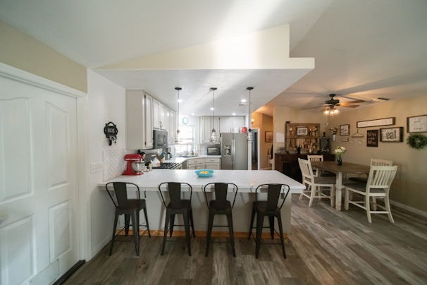 A modern kitchen and dining area with white cabinetry and a central island featuring four stools. The kitchen is equipped with stainless steel appliances, including a refrigerator and dishwasher. Overhead pendant lights illuminate the space. On the right, a wooden dining table with white chairs and a decorative setting creates a cozy ambiance. Decor on the walls and a ceiling fan complete the homely atmosphere.