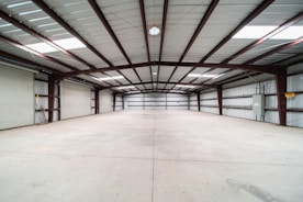 A spacious, empty warehouse featuring a concrete floor and a high metal ceiling. The structure is supported by exposed steel beams, and there are roller doors along the left side, indicating multiple points of entry. The environment is clean with evenly distributed natural light coming from skylights in the ceiling.