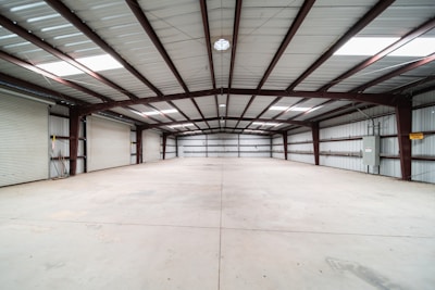 A spacious, empty warehouse featuring a concrete floor and a high metal ceiling. The structure is supported by exposed steel beams, and there are roller doors along the left side, indicating multiple points of entry. The environment is clean with evenly distributed natural light coming from skylights in the ceiling.