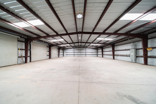 A spacious, empty warehouse featuring a concrete floor and a high metal ceiling. The structure is supported by exposed steel beams, and there are roller doors along the left side, indicating multiple points of entry. The environment is clean with evenly distributed natural light coming from skylights in the ceiling.