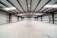 A spacious, empty warehouse featuring a concrete floor and a high metal ceiling. The structure is supported by exposed steel beams, and there are roller doors along the left side, indicating multiple points of entry. The environment is clean with evenly distributed natural light coming from skylights in the ceiling.