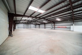 A large, empty warehouse featuring a high ceiling supported by metal beams. The walls are lined with metal panels, and the floor is a clean, smooth concrete. A partially open roller door and a sign reading 'PJ TRAILERS' are visible on one wall.