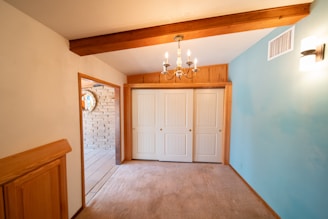 A room with beige carpet flooring, a blue accent wall, and a wooden beam on the ceiling. A chandelier is hanging from the ceiling, providing warm lighting. There is a double-door closet with white panel doors on one side and a doorway leading to a tiled area with a brick wall.