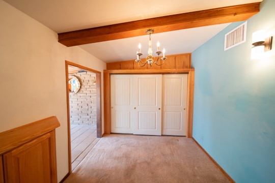 A room with beige carpet flooring, a blue accent wall, and a wooden beam on the ceiling. A chandelier is hanging from the ceiling, providing warm lighting. There is a double-door closet with white panel doors on one side and a doorway leading to a tiled area with a brick wall.