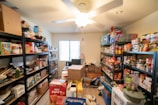 A well-stocked pantry room with metal shelves filled with various food items, canned goods, and household supplies. The shelves are organized with boxes of cereal, cans, jars, and packaged products. Cardboard boxes and household items are scattered on the floor. A ceiling fan is centrally located above, and natural light is coming through a window.