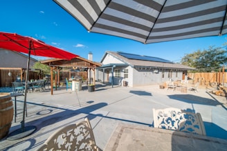 A spacious backyard features a concrete patio with various seating areas and umbrellas. There is a house with solar panels on the roof, a wooden pergola providing shade to a barbeque area, and trees along the fence line.