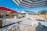 A spacious backyard features a concrete patio with various seating areas and umbrellas. There is a house with solar panels on the roof, a wooden pergola providing shade to a barbeque area, and trees along the fence line.