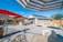 A spacious backyard features a concrete patio with various seating areas and umbrellas. There is a house with solar panels on the roof, a wooden pergola providing shade to a barbeque area, and trees along the fence line.