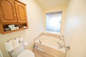 A small bathroom featuring a bathtub with modern fixtures and a frosted window. Above the toilet, a wooden cabinet with open shelves displays items like toilet paper rolls and personal care products. The walls and floor have a clean and tidy appearance, contributing to a minimalist aesthetic.