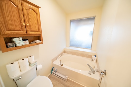 A small bathroom featuring a bathtub with modern fixtures and a frosted window. Above the toilet, a wooden cabinet with open shelves displays items like toilet paper rolls and personal care products. The walls and floor have a clean and tidy appearance, contributing to a minimalist aesthetic.