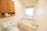 A small bathroom featuring a bathtub with modern fixtures and a frosted window. Above the toilet, a wooden cabinet with open shelves displays items like toilet paper rolls and personal care products. The walls and floor have a clean and tidy appearance, contributing to a minimalist aesthetic.