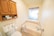 A small bathroom featuring a bathtub with modern fixtures and a frosted window. Above the toilet, a wooden cabinet with open shelves displays items like toilet paper rolls and personal care products. The walls and floor have a clean and tidy appearance, contributing to a minimalist aesthetic.