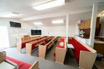 A small church interior with red cushioned pews and a pulpit at the front. Two televisions are mounted on stands, and there's a whiteboard with religious text on the wall. A ceiling with fluorescent lights provides ample illumination, and a small fan is positioned on a wooden cabinet to the right.