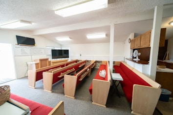 A small church interior with red cushioned pews and a pulpit at the front. Two televisions are mounted on stands, and there's a whiteboard with religious text on the wall. A ceiling with fluorescent lights provides ample illumination, and a small fan is positioned on a wooden cabinet to the right.