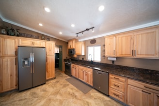 A spacious kitchen with wooden cabinetry and modern appliances, including a stainless steel refrigerator and dishwasher. The countertop is dark granite, and there is a large window above the sink allowing natural light to enter. Recessed lighting in the ceiling provides additional illumination. The flooring appears to be tile with a neutral design.