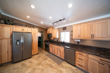 A spacious kitchen with wooden cabinetry and modern appliances, including a stainless steel refrigerator and dishwasher. The countertop is dark granite, and there is a large window above the sink allowing natural light to enter. Recessed lighting in the ceiling provides additional illumination. The flooring appears to be tile with a neutral design.
