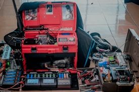 A workspace featuring various electronic and mechanical components. The central focus is a red toolbox open to reveal compartments filled with tools and electronic devices. Multiple screens display numbers, likely part of battery or voltage monitoring equipment. Nearby, a collection of small vehicles or gadgets with wheels are assembled, suggesting a hobbyist setup, possibly for robotics or model cars. Cables and hand tools are scattered about, and photos are pinned inside the toolkit lid.