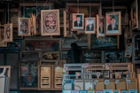 A cluttered display of various framed pictures and artworks, including portraits of men in formal attire, decorative calligraphy, and religious-themed art. The frames are ornate and predominantly golden, hanging and leaning against each other in a densely packed arrangement.