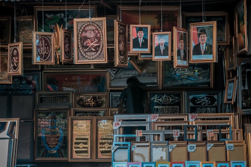 A cluttered display of various framed pictures and artworks, including portraits of men in formal attire, decorative calligraphy, and religious-themed art. The frames are ornate and predominantly golden, hanging and leaning against each other in a densely packed arrangement.