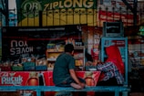 A small street-side stall selling a variety of snacks and beverages is shown, with two people seated at the counter. The stall is equipped with baskets filled with packaged goods and a small refrigerator. Brightly colored advertisements and signs hang around the stall, including one for Teh Pucuk Harum. One person is wearing a red headscarf and the other is dressed casually in a gray shirt and jeans.
