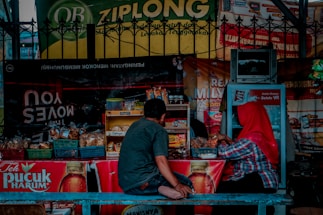 A small street-side stall selling a variety of snacks and beverages is shown, with two people seated at the counter. The stall is equipped with baskets filled with packaged goods and a small refrigerator. Brightly colored advertisements and signs hang around the stall, including one for Teh Pucuk Harum. One person is wearing a red headscarf and the other is dressed casually in a gray shirt and jeans.