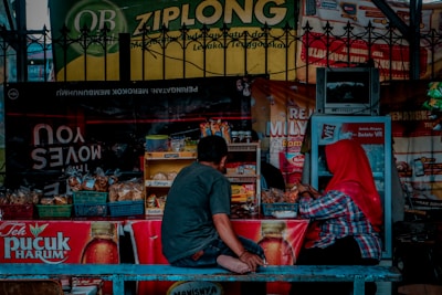 A small street-side stall selling a variety of snacks and beverages is shown, with two people seated at the counter. The stall is equipped with baskets filled with packaged goods and a small refrigerator. Brightly colored advertisements and signs hang around the stall, including one for Teh Pucuk Harum. One person is wearing a red headscarf and the other is dressed casually in a gray shirt and jeans.