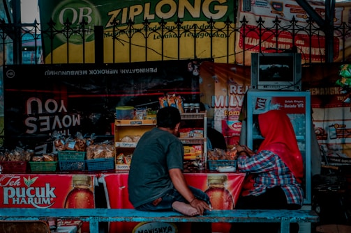A small street-side stall selling a variety of snacks and beverages is shown, with two people seated at the counter. The stall is equipped with baskets filled with packaged goods and a small refrigerator. Brightly colored advertisements and signs hang around the stall, including one for Teh Pucuk Harum. One person is wearing a red headscarf and the other is dressed casually in a gray shirt and jeans.