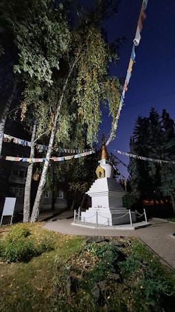 A religious monument with a golden top is set in a well-maintained garden. The area is decorated with colorful prayer flags hanging between tall trees. The scene is illuminated in a manner that suggests it is either early morning or late evening, giving the surroundings a serene and solemn atmosphere.