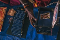 A person's hand interacts with a stack of blue booklets and a small blue box with an image on the lid. Nearby, there are some currency notes and a decorative red cover with yellow and golden accents. The scene is set against a dark blue surface.