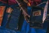 A person's hand interacts with a stack of blue booklets and a small blue box with an image on the lid. Nearby, there are some currency notes and a decorative red cover with yellow and golden accents. The scene is set against a dark blue surface.