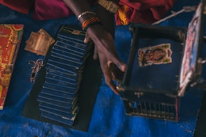 A person's hand interacts with a stack of blue booklets and a small blue box with an image on the lid. Nearby, there are some currency notes and a decorative red cover with yellow and golden accents. The scene is set against a dark blue surface.