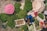 An overhead view of a marketplace scene with an assortment of greenery, flowers, and produce. A person is sitting among large piles of herbs and sacks filled with leaves. Pink flowers are arranged prominently, and there are containers with lotus buds. The scene is vibrant and earthy, with a focus on natural produce.