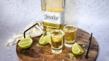 A wooden surface holds a bottle of tequila labeled 'El Jimador' alongside two filled shot glasses with salted rims. Lime wedges are placed near the glasses, and a metal stirrer rests on the board. A light-colored cloth partially drapes the background.