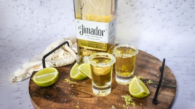 A wooden surface holds a bottle of tequila labeled 'El Jimador' alongside two filled shot glasses with salted rims. Lime wedges are placed near the glasses, and a metal stirrer rests on the board. A light-colored cloth partially drapes the background.