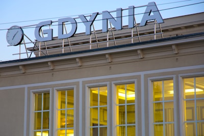 An architectural detail of a building with large rectangular windows illuminated by warm yellow light. Above the building, there is a large sign with the word 'GDYNIA' in bold letters and a round clock on display, all set against a soft blue sky.