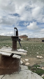A rustic water pump stands in the foreground, surrounded by a stone base and wooden planks. In the background, an old house with rustic walls and a slanted roof is visible under a cloudy sky. The ground is scattered with rocks and sparse patches of grass.