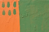 A textured wall is painted in two contrasting colors: a vibrant orange on the left and a muted green on the right. The orange section includes six oval green paint marks arranged in two vertical columns.