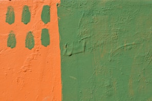 A textured wall is painted in two contrasting colors: a vibrant orange on the left and a muted green on the right. The orange section includes six oval green paint marks arranged in two vertical columns.